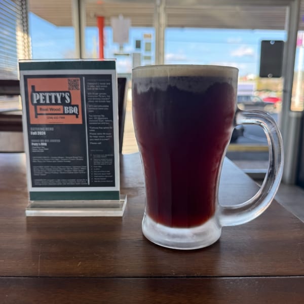 Dark beverage in a glass mug and a Petty's BBQ catering menu on a wooden table.