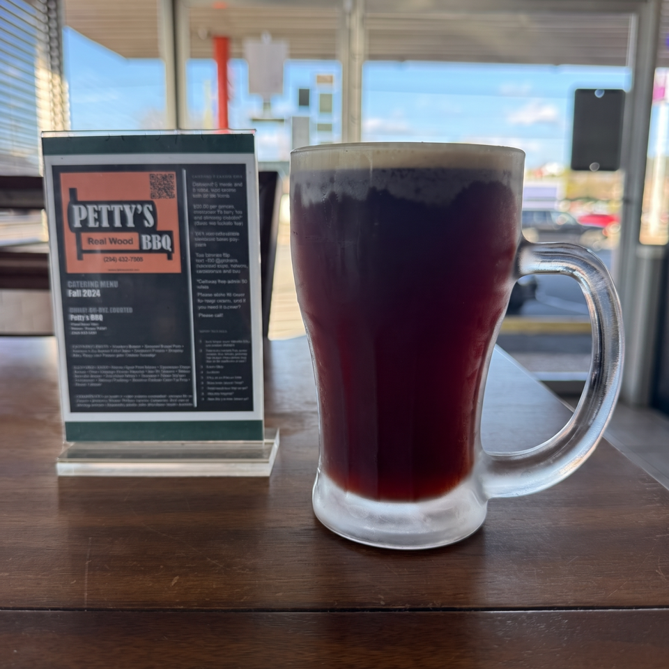 Dark beverage in a glass mug and a Petty's BBQ catering menu on a wooden table.