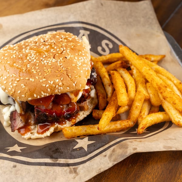 Brisket sandwich with BBQ sauce and white sauce, served with seasoned fries on branded paper.