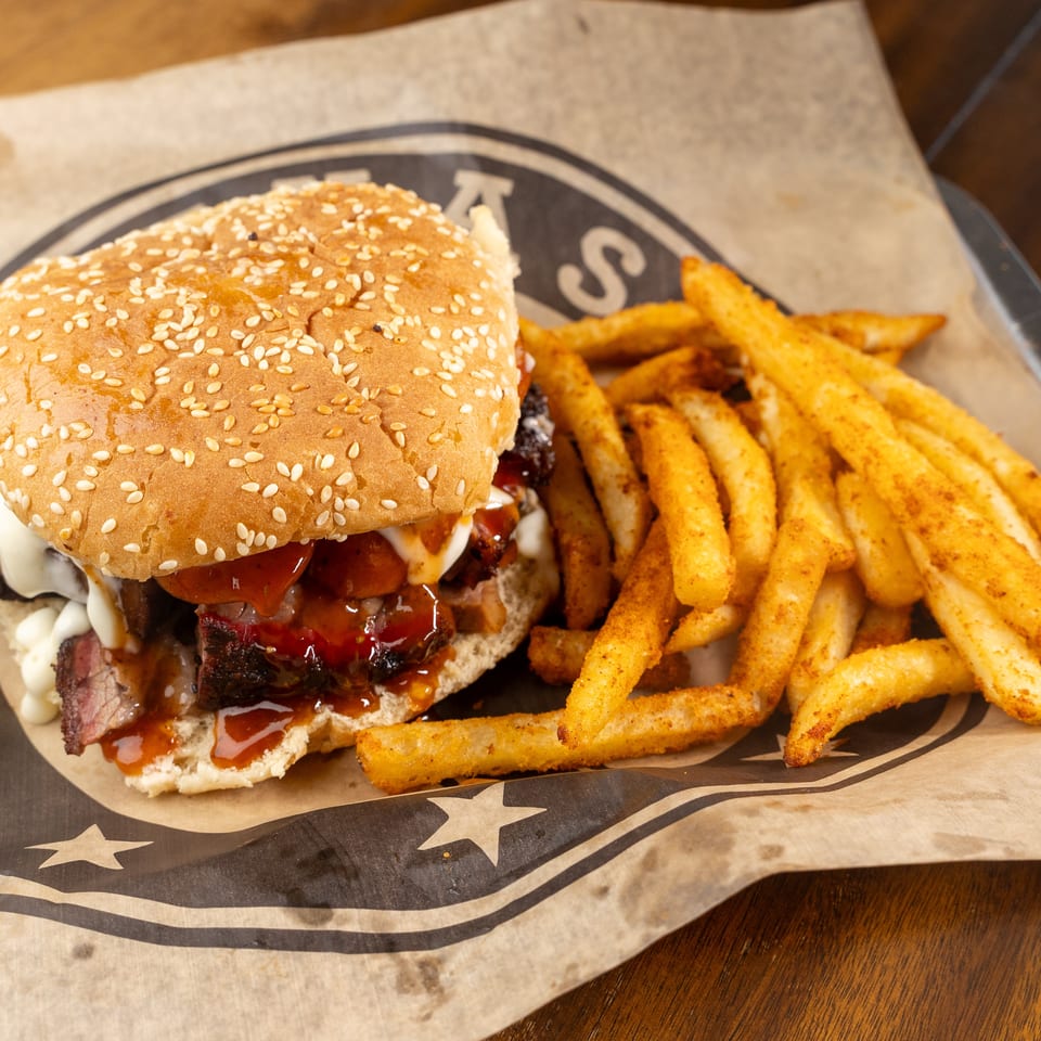 Brisket sandwich with BBQ sauce and white sauce, served with seasoned fries on branded paper.