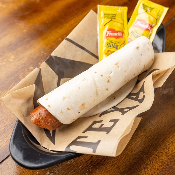 A tortilla-wrapped hot dog in a basket with two French's mustard packets on a wooden table.