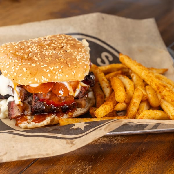 Brisket sandwich with barbecue sauce and mayo, served with seasoned fries on branded paper.