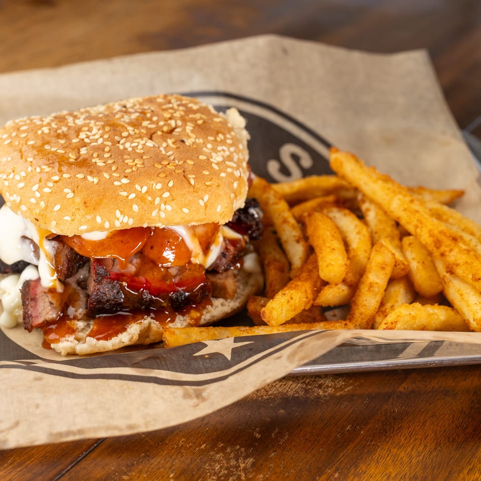Brisket sandwich with barbecue sauce and mayo, served with seasoned fries on branded paper.